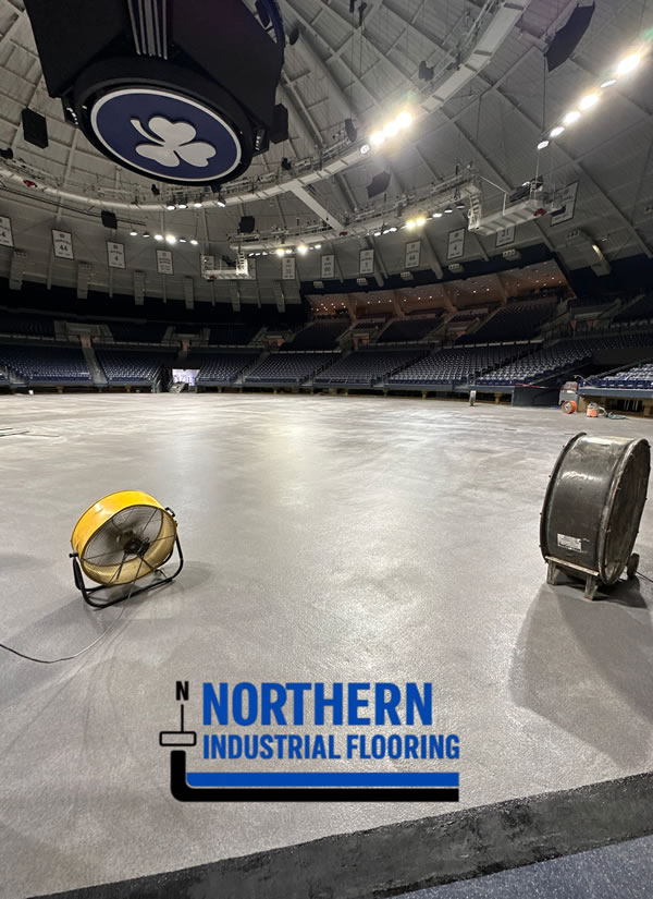 Arena with a freshly installed concrete floor under construction; orange/yellow work fan and a large cable reel on the floor, logo reads 'Northern Industrial Flooring' in the foreground.